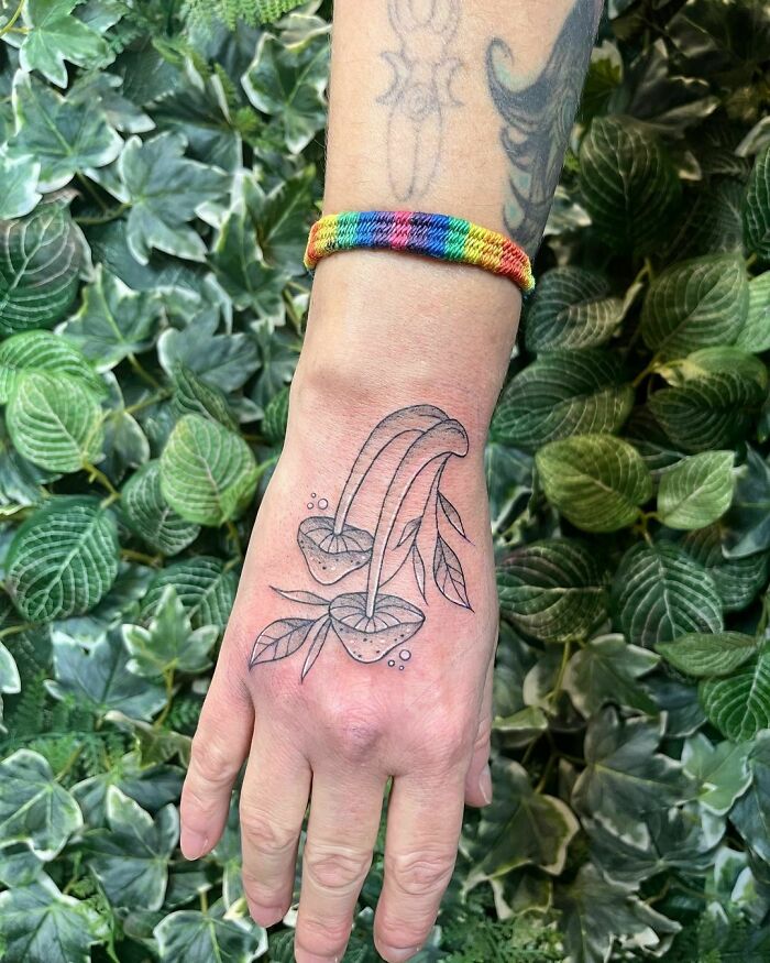 Hand with nature tattoo of plants, wearing a rainbow bracelet, surrounded by green leaves.