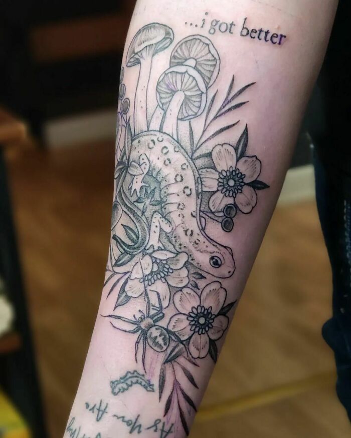 Nature tattoo featuring a salamander, mushrooms, and flowers on forearm, with text "I got better" above the design.