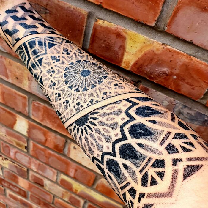 Arm tattoo featuring intricate geometric designs and patterns, highlighting shapes and forms on a brick wall background.