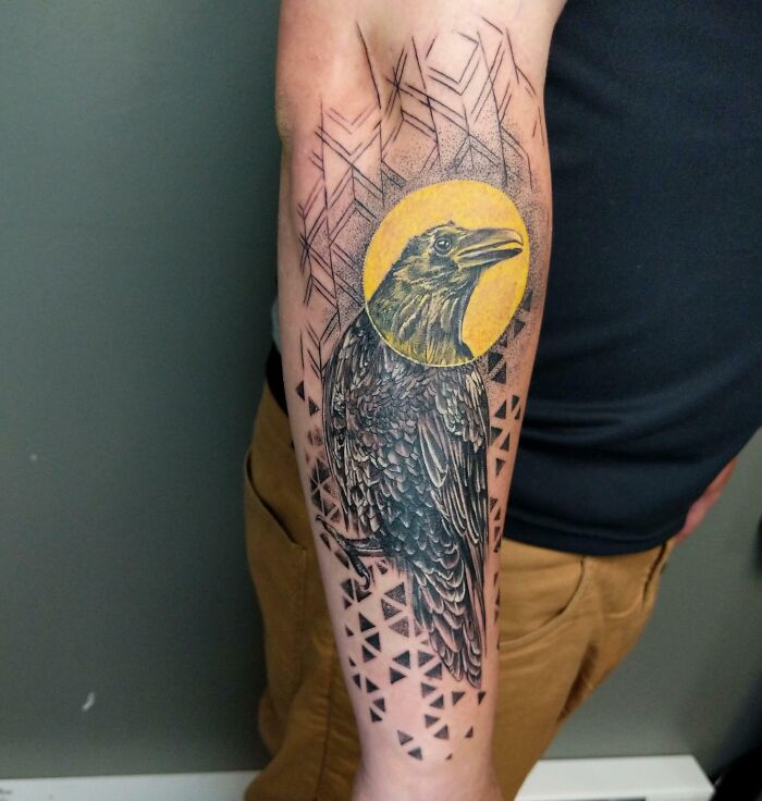 Geometric tattoo design featuring a detailed raven with a circular yellow background and triangular patterns on the forearm.