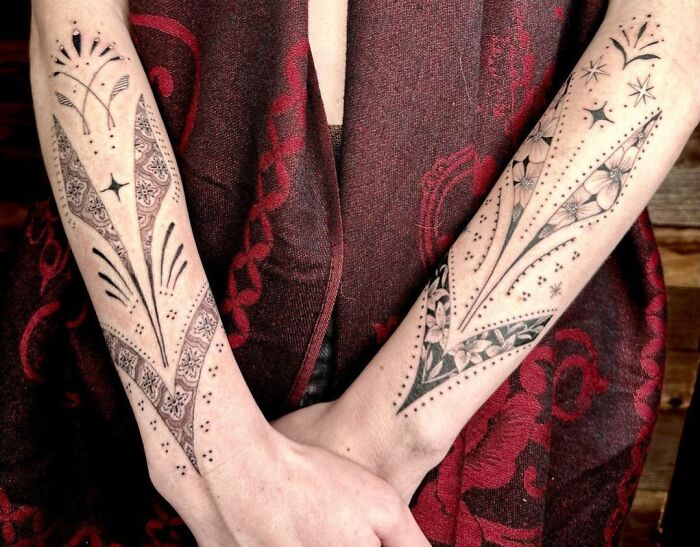 Geometric tattoos on forearms, featuring intricate patterns and designs against a red and purple fabric background.