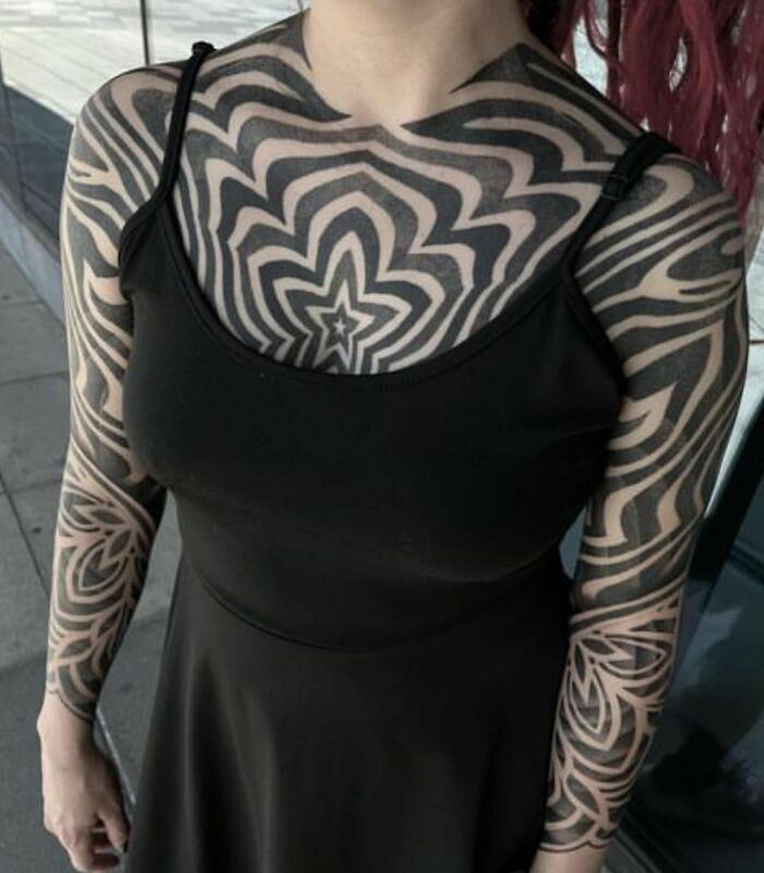 Person with intricate geometric tattoos covering chest and arms, wearing a black sleeveless top.