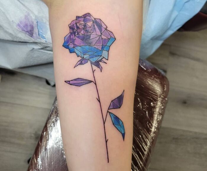 Geometric tattoo of a blue rose on a forearm, featuring angular shapes and a detailed stem.