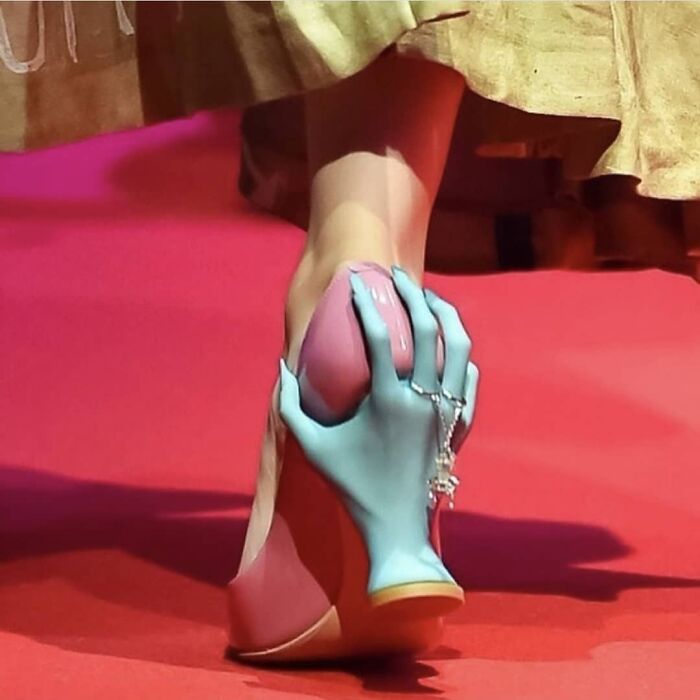 Weird shoes with a blue hand-shaped heel holding a pink platform on a red carpet.