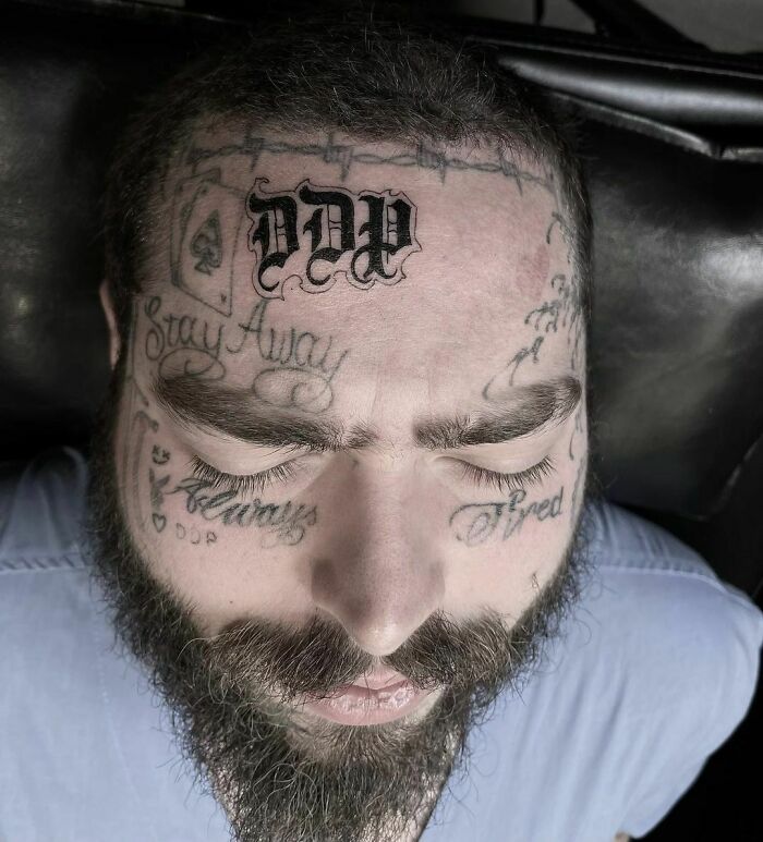 Celebrity with multiple face tattoos, including "Stay Away" and "Tired," lying on a black surface, eyes closed.