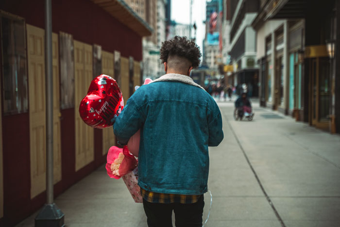 Man in denim jacket and black pants holding red heart shaped balloon and flowers