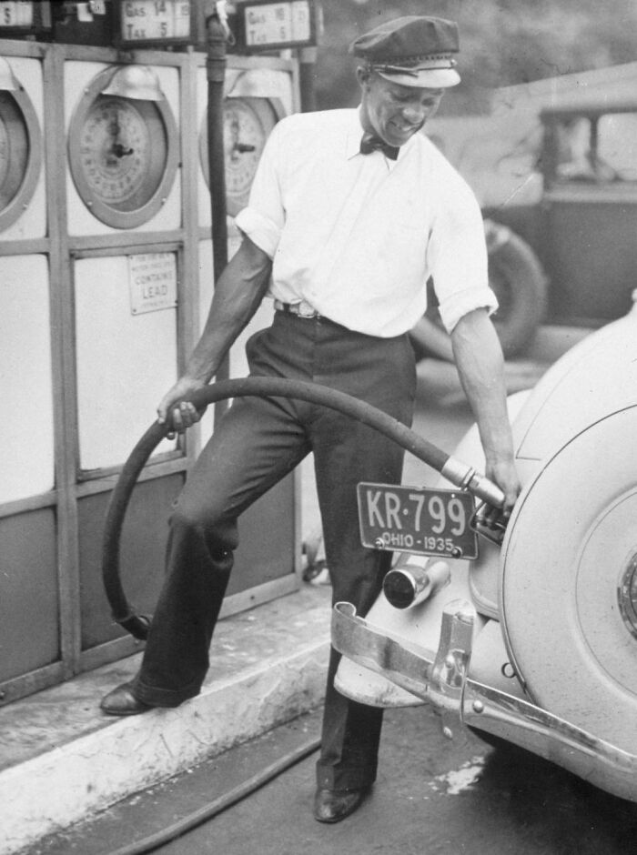 El atleta estadounidense Jesse Owens llena el depósito de un coche en una gasolinera con su uniforme de gorra, camisa y pajarita. Owens trabajó como empleado de gasolinera para ayudar a financiar sus estudios en la Universidad Estatal de Ohio. (Estados Unidos, 1935)