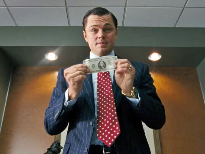 Actor holding a dollar bill, dressed in a suit and red polka dot tie, symbolizing highest-paid actors in entertainment.