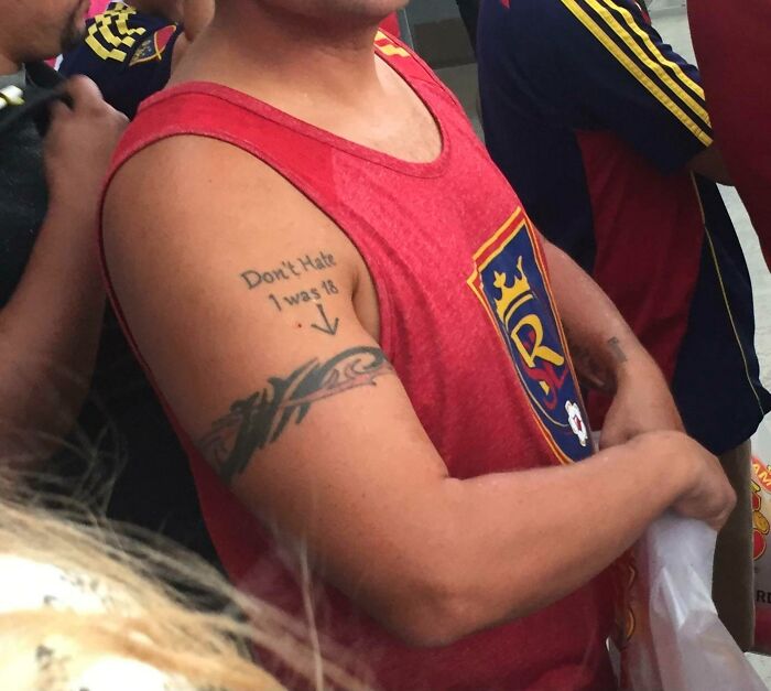Man with funny tattoo reading "Don't Hate, I was 18" on his arm, wearing a red sports tank top at an event.