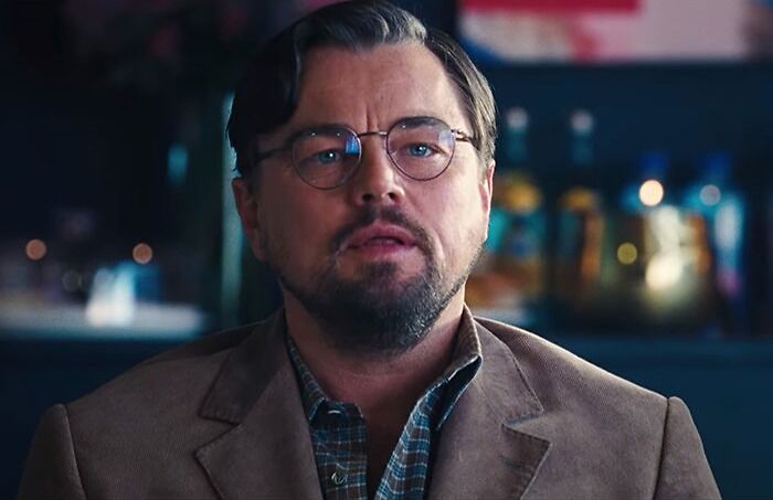 Male highest-paid actor with glasses and beard wearing a brown blazer in a dimly lit indoor setting