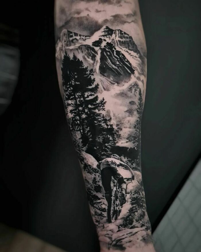 Realistic tattoo of a mountain biker on a trail with mountains and trees in black and gray tones.