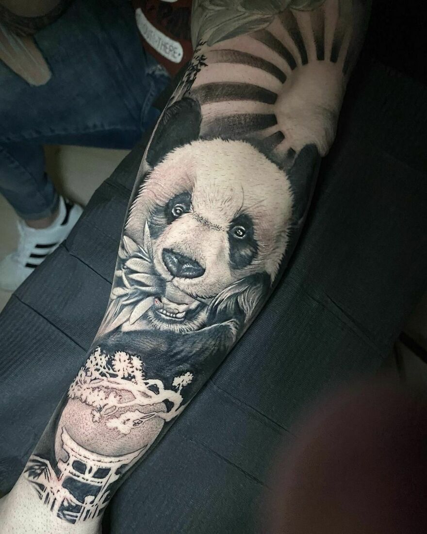 Realistic tattoo of a panda eating bamboo with a sun design above its head on an arm.