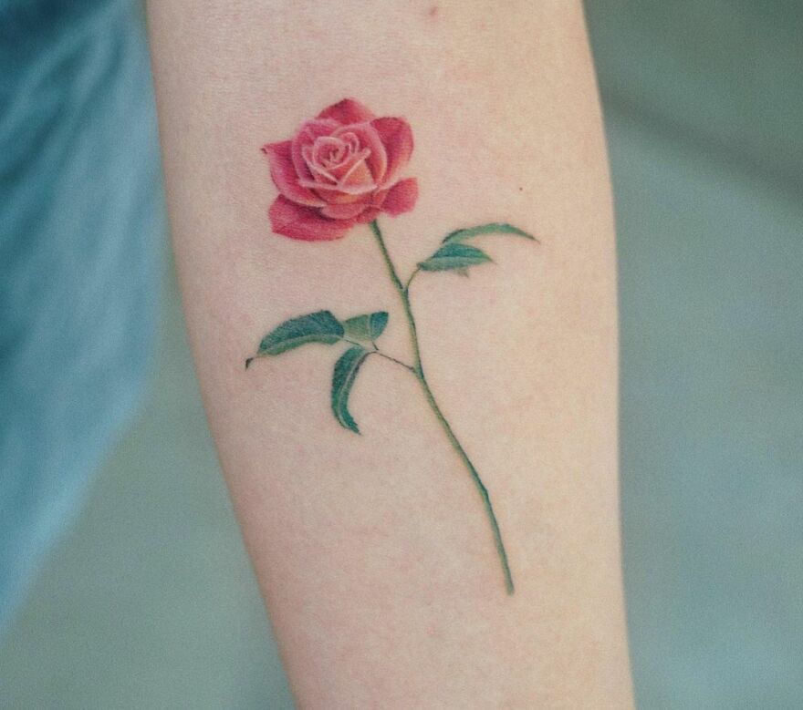 A super realistic tattoo of a pink rose with a green stem and leaves on a person's forearm.