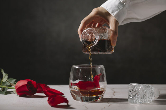 A hand pouring brandy from carafe to glass with red rose petals