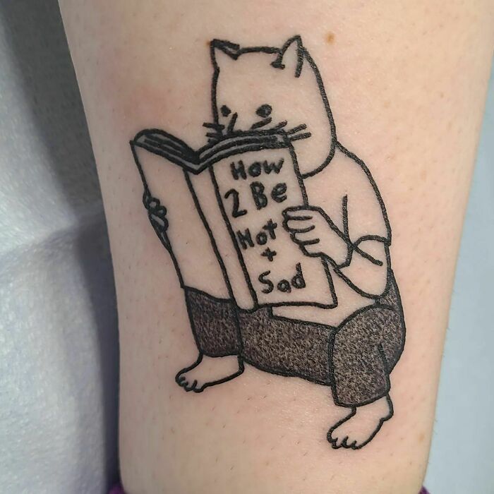Cat reading a book titled "How 2 Be Not Sad" tattoo, showcasing self-love through humor and minimalist design.