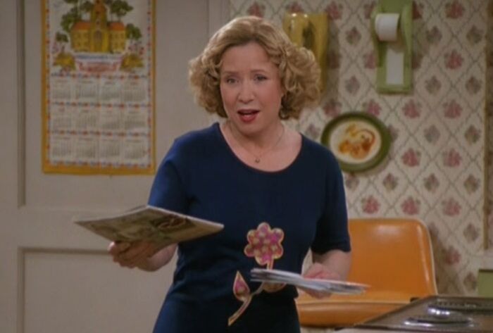 Kitty Forman is wearing a dark blue dress, holding newspapers, and letters, and talking