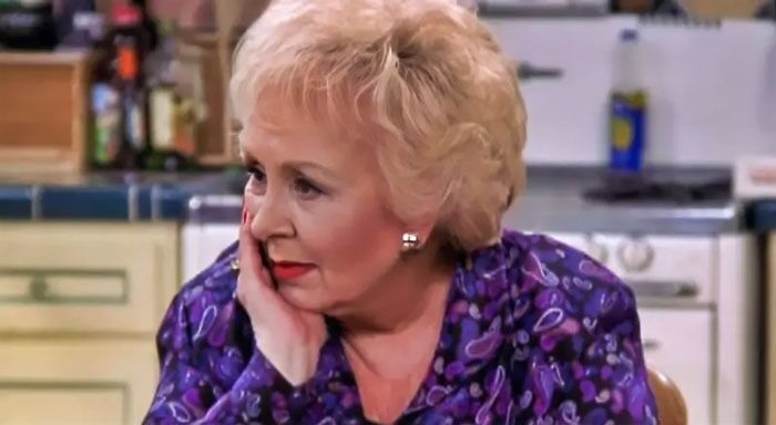 Marie Barone in a purple blouse is holding her cheek and sitting in the kitchen