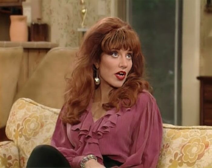Peg Bundy in a bordo blouse is sitting on the sofa and talking