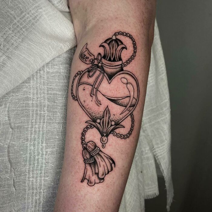 Heart-shaped self-love tattoo with intricate design and tassel on arm, celebrating positivity and personal empowerment.