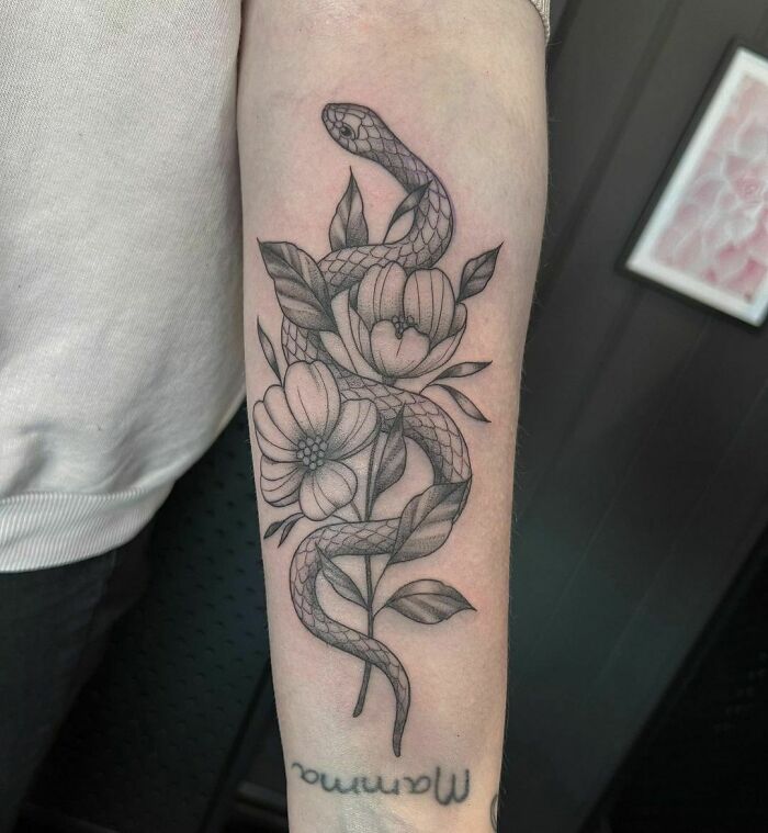 Snake with flower tattoo on forearm, showcasing elegance and beauty in a detailed black and white design.