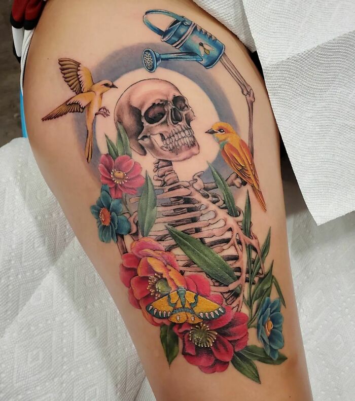 Intricate self-love tattoo with a detailed skeleton, colorful flowers, birds, and a watering can, symbolizing growth and transformation.