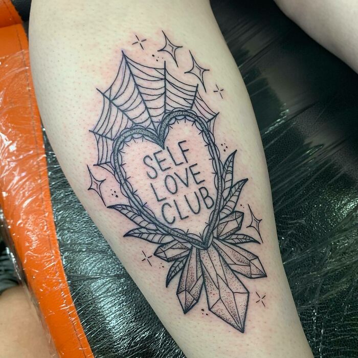 Heart-shaped self-love tattoo with "Self Love Club" text, surrounded by crystals and web design, on a person's leg.