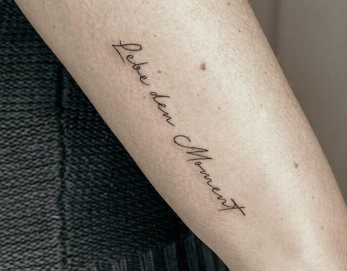 A delicate script tattoo on a forearm reads "Lebe den Moment," embodying self-love and empowerment.