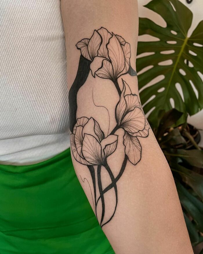 Elegant flower tattoo in black ink on forearm, showcasing intricate petal details with a plant in the background.