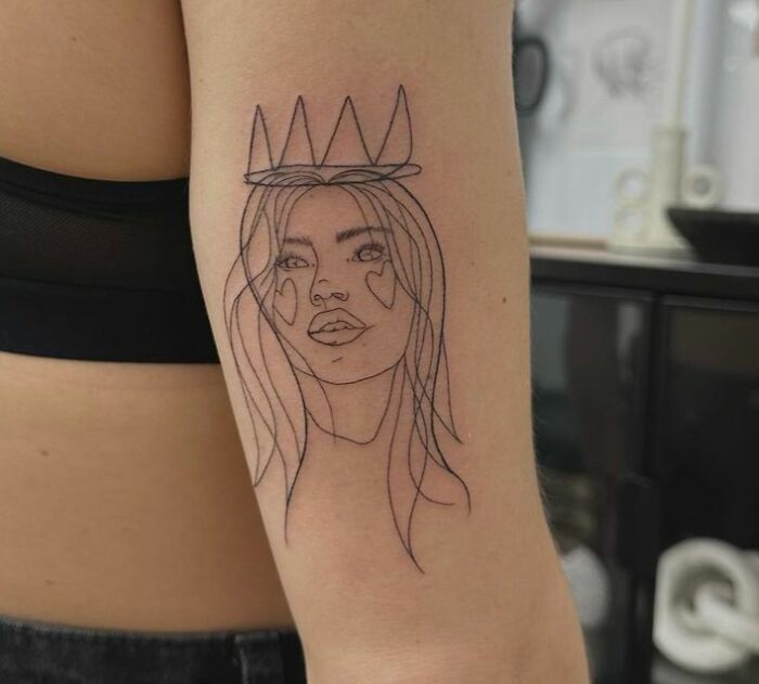 Minimalist self-love tattoo of a crowned woman with heart-shaped cheeks on an arm.