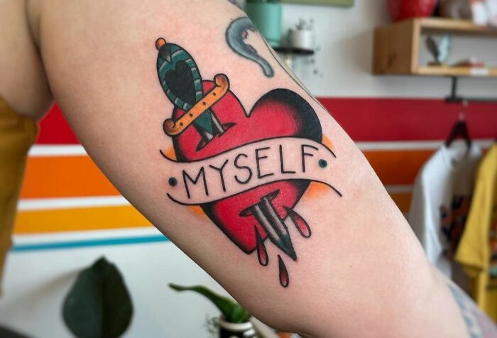 Self-love tattoo with a heart and dagger, reading "Myself," on a forearm, symbolizing personal empowerment.