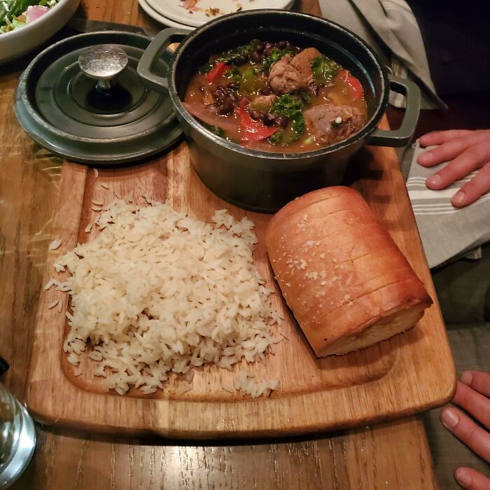 Brazilian Stew (Feijoada) In A Mini Dutch Oven Served With Rice & Bread On A Chopping Board