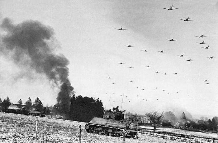 Low Flying C-47 Transport Planes Roar Overhead As They Carry Supplies To The Besieged American Forces Battling The Germans At Bastogne, During The Enemy Breakthrough On January 6, 1945 In Belgium. In The Distance, Smoke Rises From Wrecked German Equipment, While In The Foreground, American Tanks Move Up To Support The Infantry In The Fighting