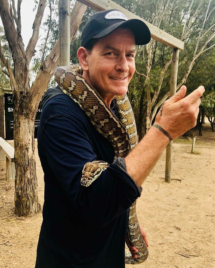 Celebrity with a snake draped over their shoulders, wearing a cap and black shirt, smiling outdoors.