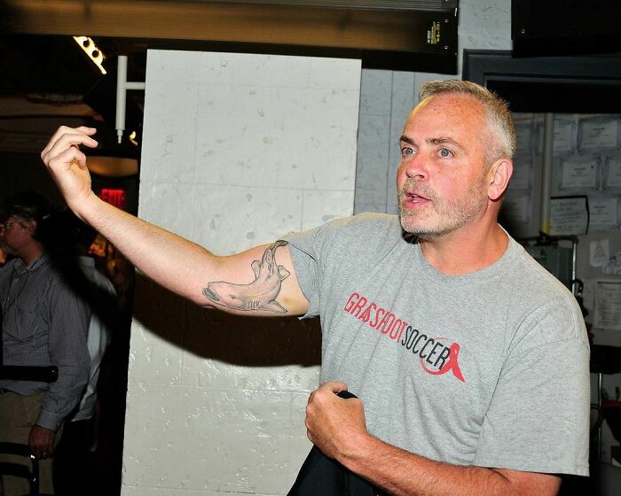Man with a tattoo on his arm gestures energetically, wearing a "Grassroot Soccer" t-shirt.