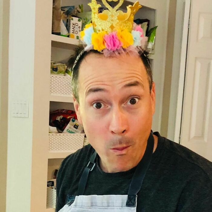 A man with a surprised expression wearing a playful crown, standing in a kitchen.