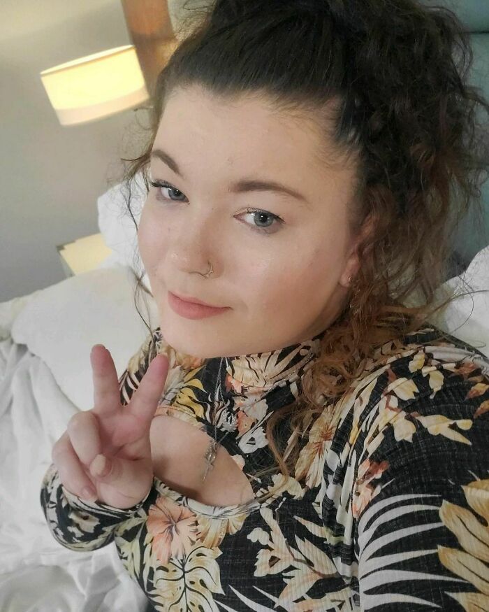 Young woman with curled hair and a nose ring, wearing a floral top, making a peace sign in a cozy room; concept of celebrities.
