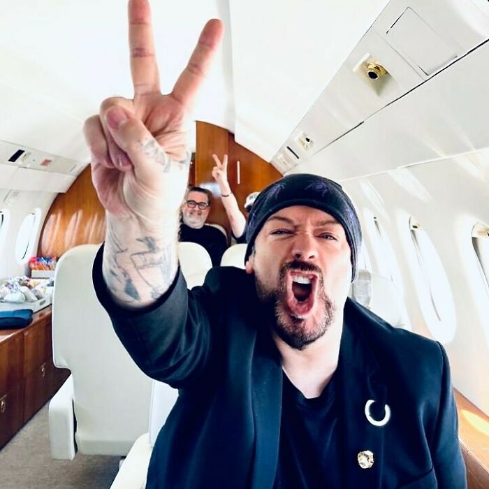 Man wearing a black beanie and blazer inside a private jet, gesturing peace signs with expressive face.