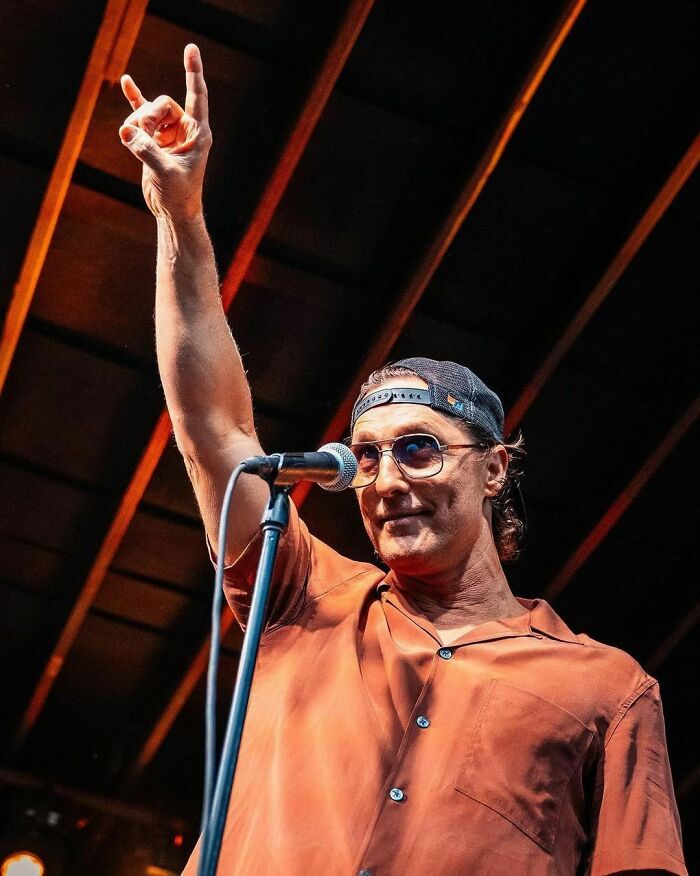 Celebrity wearing sunglasses and a cap, raising a hand in front of a microphone, under stage lights.