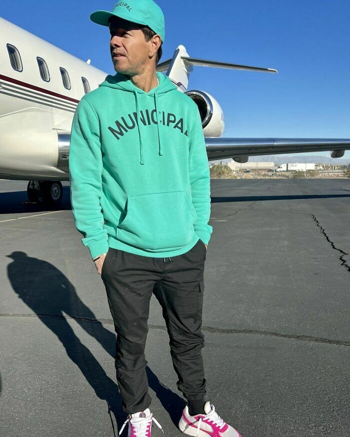 Man in a teal hoodie and cap stands by a private jet on a sunny day. Celebrities and jail theme.
