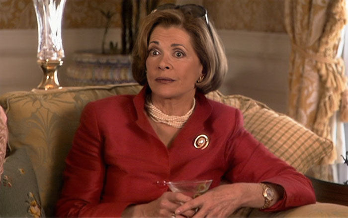 Lucille Bluth in a red top, holding a cocktail, sitting on a sofa and is looking surprised