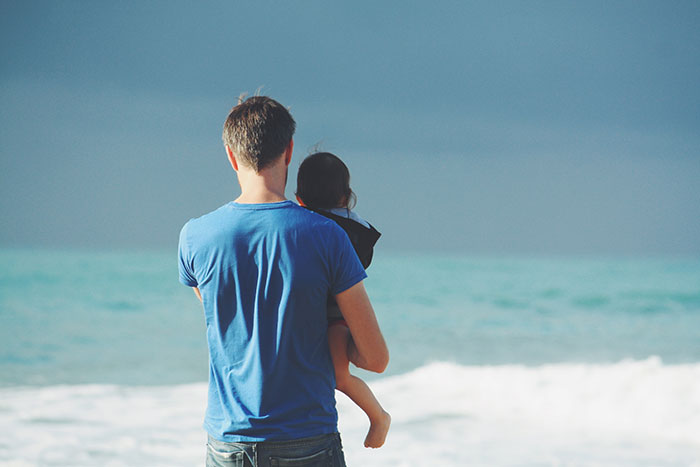 Man wears blue t-shirt holding child near ocean under blue sky at daytime