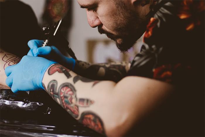 Tattoo artist inking a client's arm, showing detailed tattoos and wearing blue gloves. Tattoo artist inking a client's arm, showing detailed tattoos and wearing blue gloves.