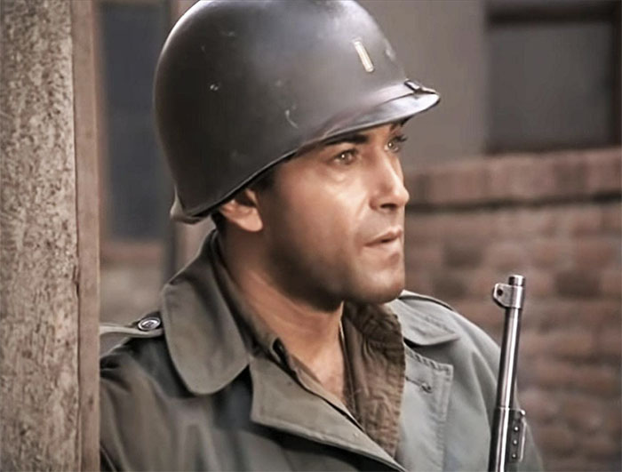Rick Jason standing near a wall with a helmet on and holding a gun 