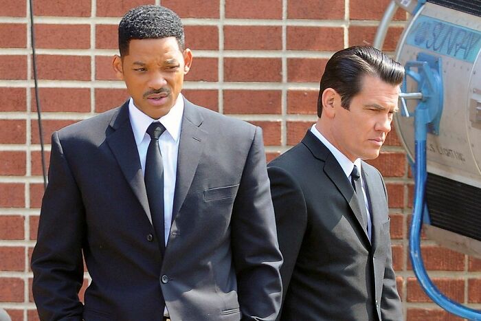 Two highest-paid actors in black suits standing outside against a brick wall, portraying serious expressions in a film scene.