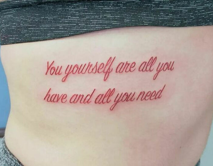 Tattoo with empowering self-love quote: "You yourself are all you have and all you need," in red script font on skin.