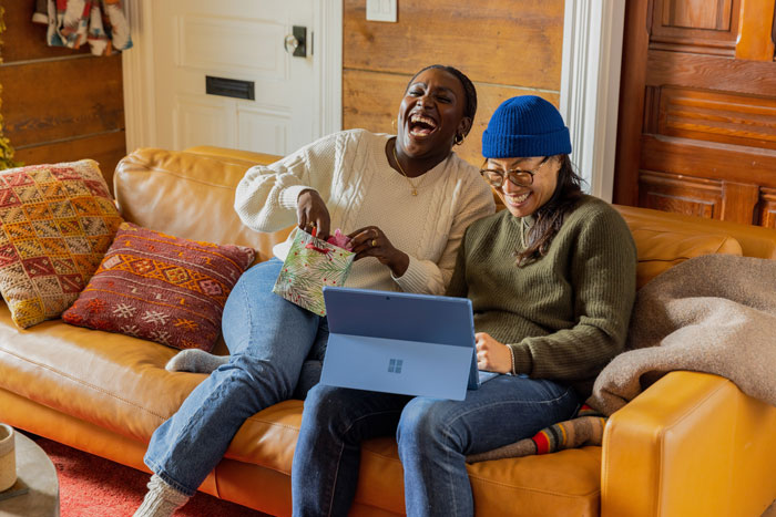 Two friends are seated on a yellow sofa, watching a laptop and laughing together
