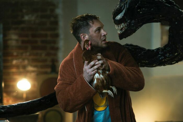 Actor Tom Hardy holding a chicken while facing a dark alien-like creature in a suspenseful movie scene.
