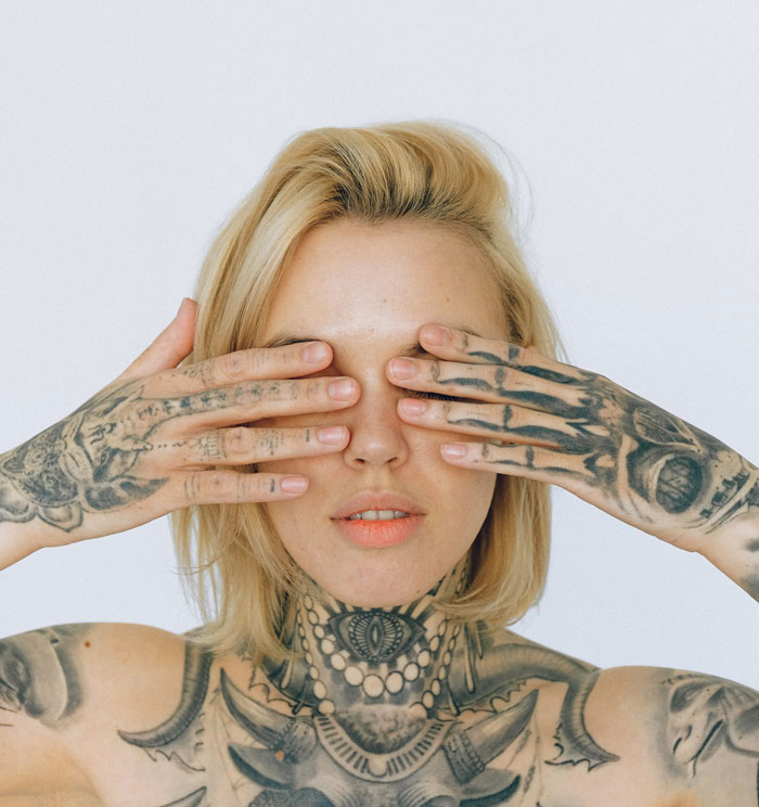 A woman with tattoos covers her eyes, showcasing intricate designs on her hands and neck.