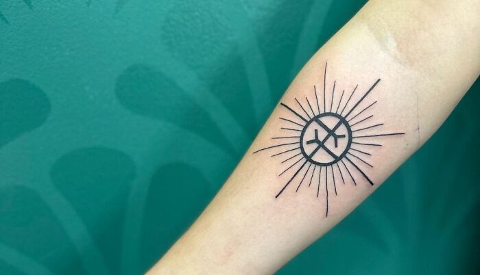 Geometric tattoo of a sun with rays and circular patterns on forearm against a green background.