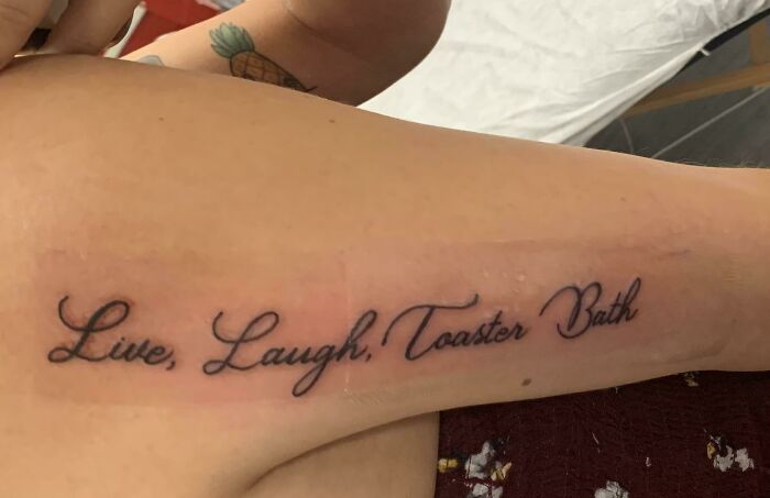 Elegant script tattoo saying "Live, Laugh, Toaster Bath" on forearm.